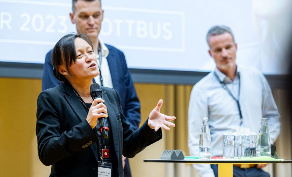 Dr. Roh Pin Lee gestikuliert auf der Bühne, im Hintergrund stehen Sebastian Kießling und Rico Schneider