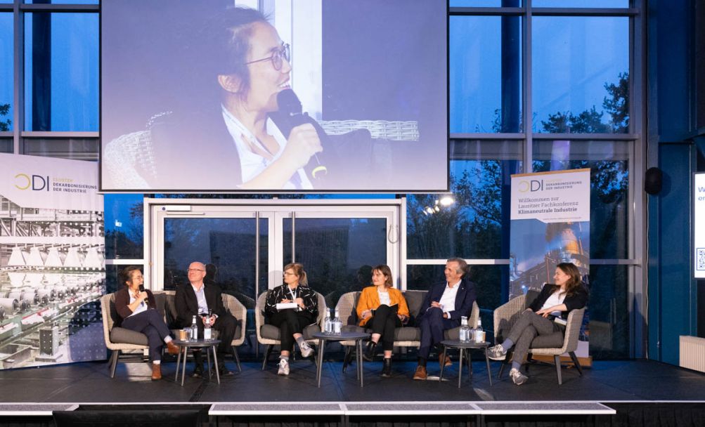 Sechs Personen sitzend unterhalten sich auf einer Bühne nebeneinander, hängende Leinwand im Hintergrund