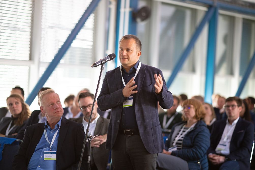 Mann vor Mikrofon mit Publikum im Hintergrund